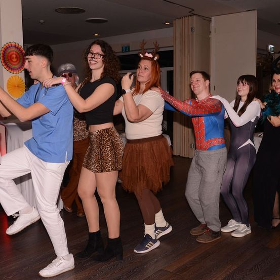 Eine Gruppe von Menschen tanzt zusammen in einem Raum, einige sind in Kostümen gekleidet. Ein Mann in einem Spider-Man-Kostüm und eine Frau mit einer Rehkopfbedeckung sind darunter.