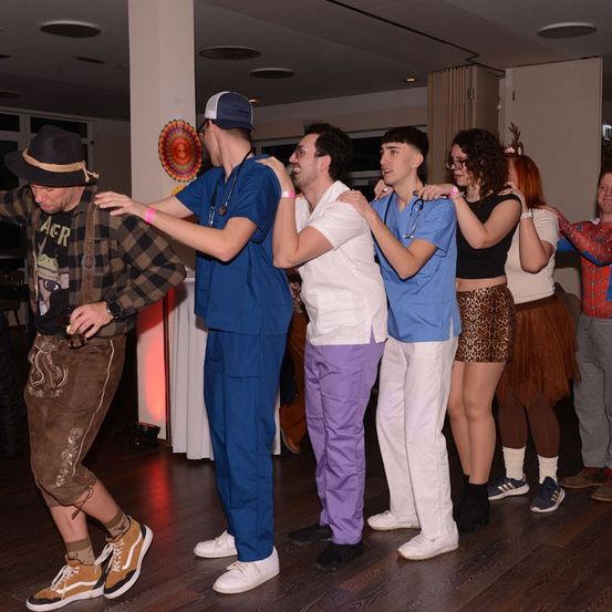 Eine Gruppe von Menschen in verschiedenen Kostümen tanzt in einem Raum, wobei zwei Männer blaue Scrubs tragen und einer ein Stethoskop hält.