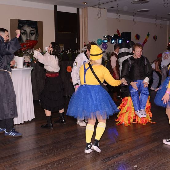 Eine Gruppe von Menschen in verschiedenen Kostümen tanzt auf einem Holzboden. Eine Frau in einer blauen Tüll- und gelben Oberbekleidung sticht hervor. Andere tragen schwarze, graue und bunte Outfits. Ein Tisch mit Getränken und ein großer Bilderrahmen sind im Hintergrund.