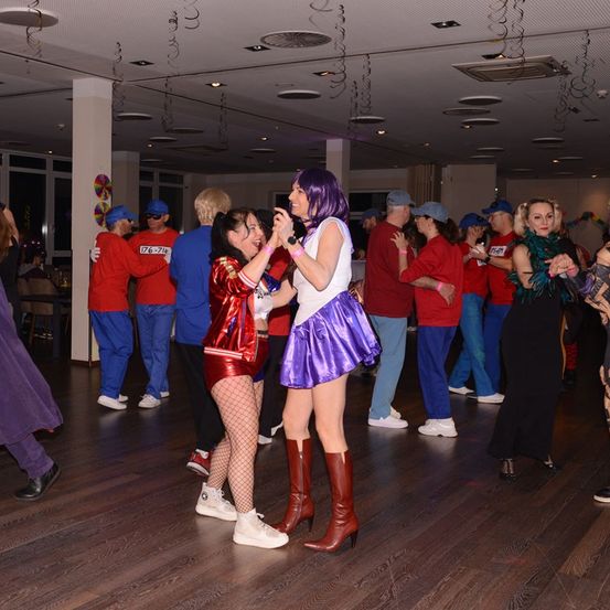 Eine Gruppe von Menschen in verschiedenen Kostümen tanzt in einem Raum. Zwei Frauen vor ihnen singen und lächeln. Sie haben verschiedenfarbige Perücken und Stiefel. Hinter ihnen stehen Menschen in passenden roten und blauen Kostümen. Der Raum hat einen Holzboden und weiße Wände.