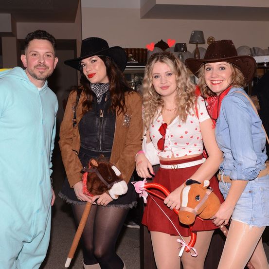 Vier Personen in Cowboy- und Cowgirl-Kleidung, lächelnd für ein Foto. Eine Frau hält ein Spielzeugpferd, eine andere hat ein Band. Ein Mann in Hellblau und eine Frau in Jeans vervollständigen die Gruppe.