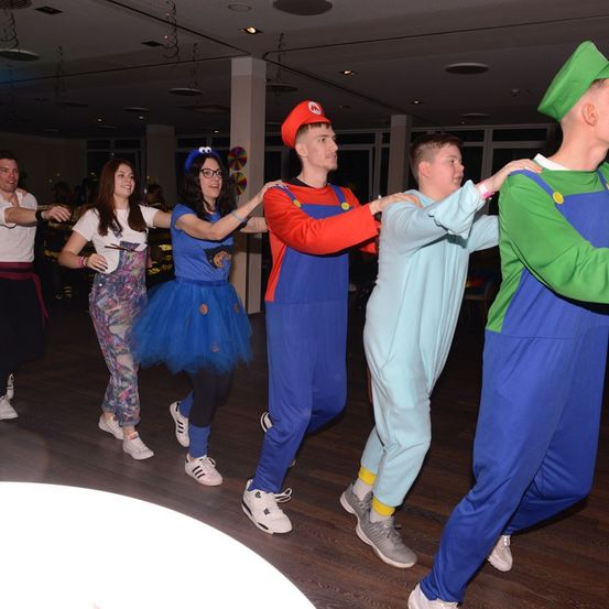 Eine Gruppe von Menschen in Nintendo-Kostümen tanzt auf einer Party. Die Charaktere umfassen Mario, Luigi und andere. Sie tragen passende Kostüme und Turnschuhe.