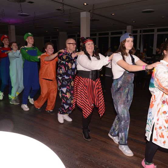 Eine Gruppe von Menschen in verschiedenen Kostümen tanzt gemeinsam auf einem Holzboden, wobei eine Frau in einem schwarz-roten Kleid den Tanz anführt.