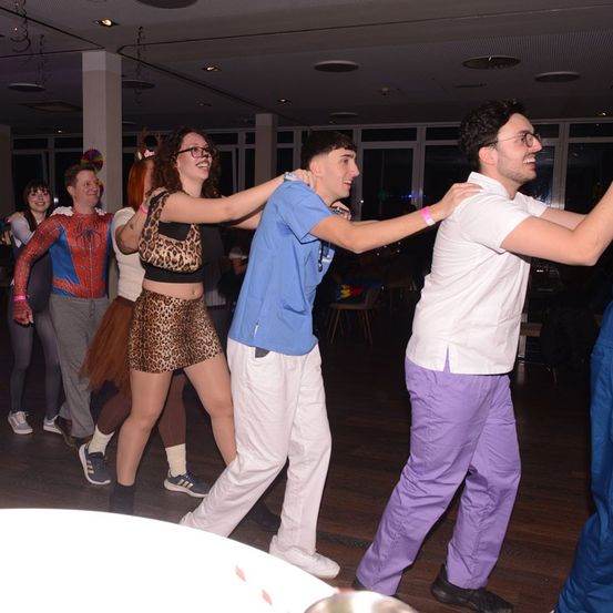 Eine Gruppe von Menschen tanzt auf einem Holzboden. Eine Frau in einem Leopardenrock und einem blauen Shirt steht in der Mitte. Zwei Männer sind auf beiden Seiten von ihr. Einige Leute stehen hinter ihnen. Der Boden ist mit warmer Beleuchtung erleuchtet.
