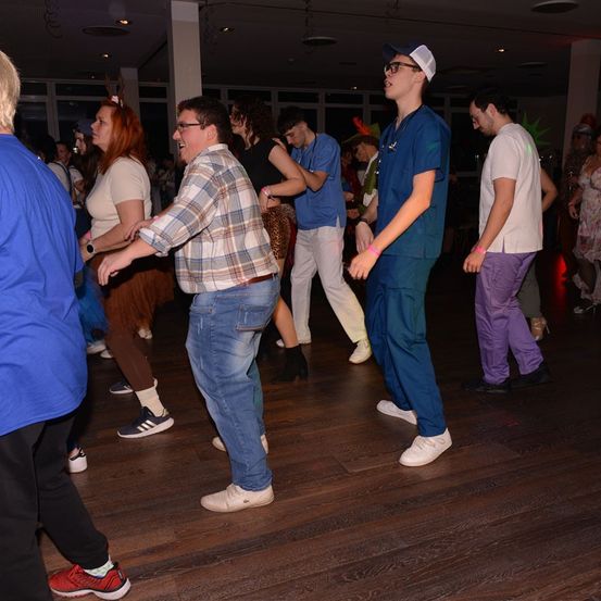 Eine Gruppe von Menschen in verschiedenen Kostümen tanzt auf einem Holzboden. Ein Mann in einem karierten Hemd lächelt. Ein Mann in Scrubs hält ein Stethoskop. Ein Mann in lila Hose trägt ein weißes Hemd.