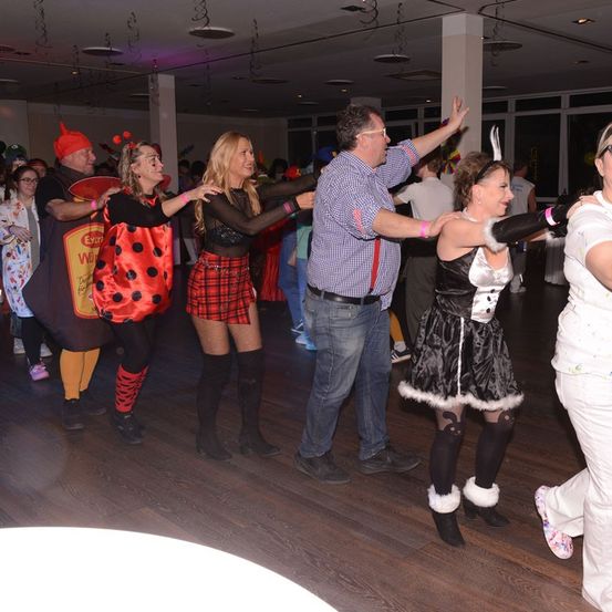 Menschen tanzen in einem Raum, gekleidet in verschiedenen Kostümen, mit einer Person in einem Marienkäfer-Outfit und einer anderen in einer Hexen-Verkleidung. Der Raum hat Holzböden und weiße Wände.