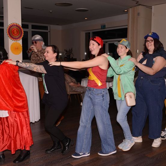 Fünf Frauen in verschiedenen Kostümen tanzen zusammen auf einem Holzboden, eine trägt einen roten Umhang und eine andere einen Hut. Ein Mann trinkt im Hintergrund ein Getränk.