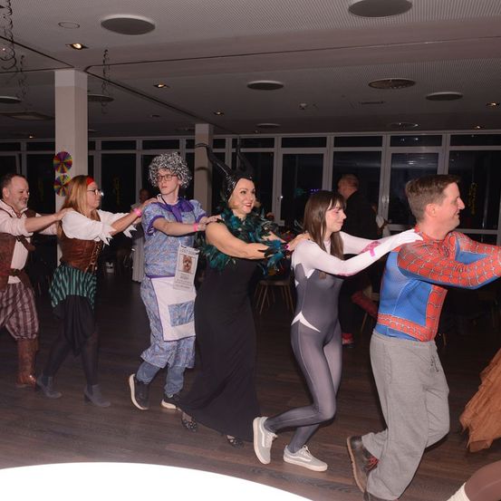 Eine Gruppe von Menschen tanzt in einem Saal. Sie sind alle in Kostümen gekleidet. Einige tragen Brillen und einer trägt eine Kopfbedeckung. Der Raum hat Holzböden und Glaswände.