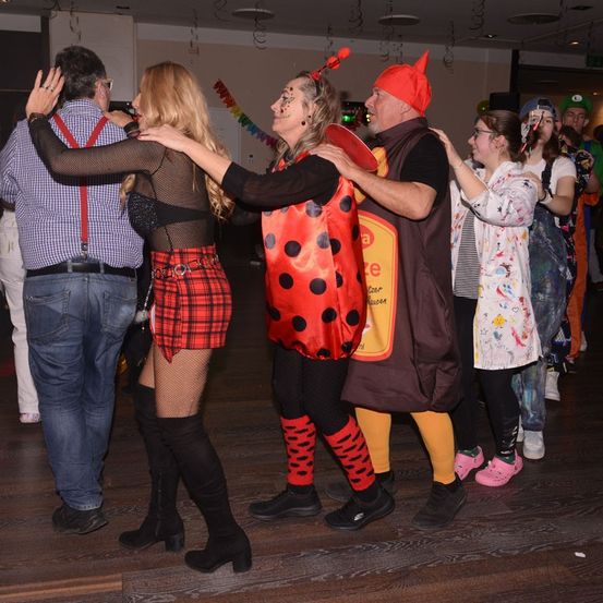Mehrere Menschen in Kostümen tanzen in einem Saal. Eine Frau in einem Marienkäfer-Kostüm wird von einem Mann in einem Schokoladensaucen-Kostüm gehalten.