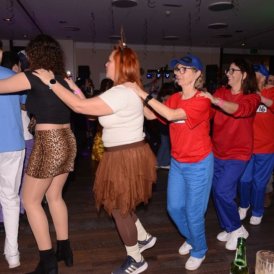 Eine Gruppe von Frauen in roten und blauen Hosen tanzt auf einer Party zusammen. Die Frau in Weiß hält die Frau in Schwarz fest.