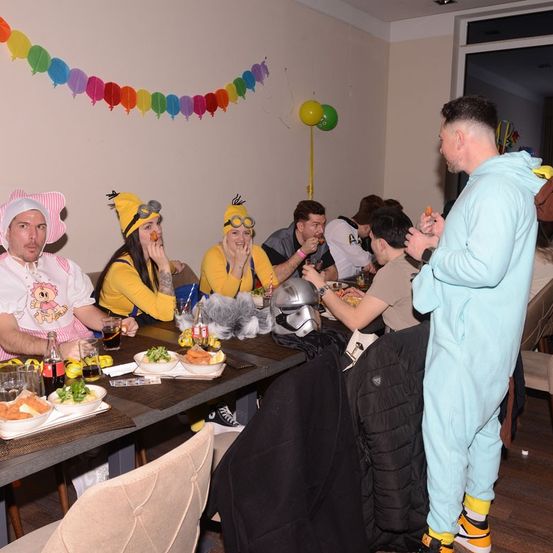 Eine Gruppe von Menschen in Kostümen, die an einem Tisch mit Essen und Getränken sitzen. Der Mann im blauen Kostüm steht neben einer schwarzen Jacke.