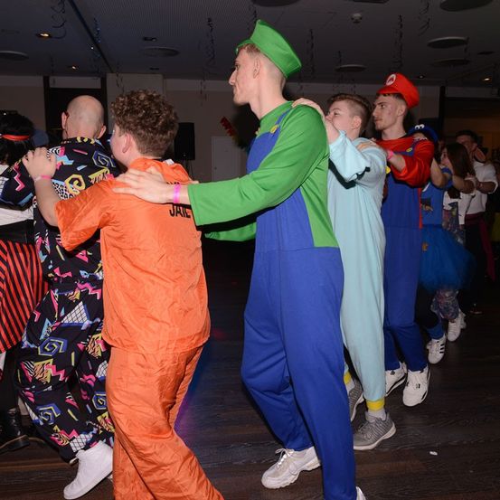 Mehrere Leute tanzen auf einem Holzboden und tragen Kostüme. Eine Person ist als Mario verkleidet, eine andere als Luigi. Einige tragen bunte Jumpsuits. An der Wand steht ein Lautsprecher.