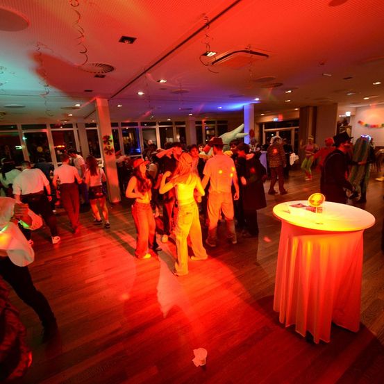 Eine Gruppe von Menschen tanzt auf einer Tanzfläche mit roten Lichtern, einige in Kostümen. Ein Tisch mit einer Kugel darauf steht in der Nähe der Wand.