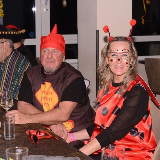 Zwei Personen in Kostümen sitzen an einem Tisch. Der Mann trägt einen roten Kegelhut und ein Schild mit der Aufschrift 'Et tich nken ann.' Die Frau hat einen Marienkäfer-Gesichtsaufkleber und ein rotes Kleid. Hinter ihnen ist ein Fenster.