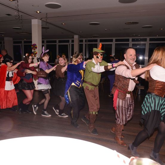 Eine Gruppe von Menschen tanzt in einem Saal in verschiedenen Kostümen, darunter Hüte, Stiefel und Masken. Der Boden ist aus Holz und die Wände haben Glasfenster.