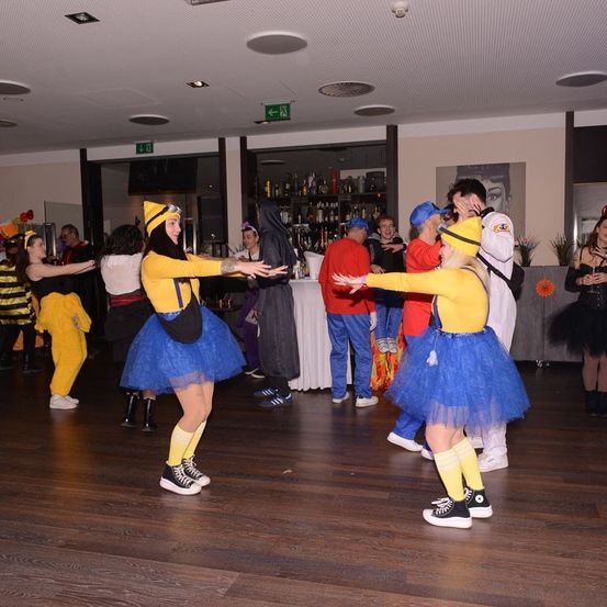 Eine Gruppe von Menschen in Kostümen, darunter Minions und eine Biene, tanzt in einem Saal. Ein Barbereich ist im Hintergrund sichtbar.