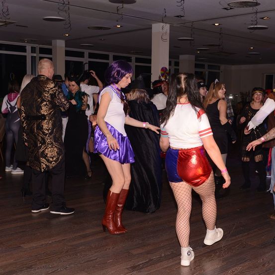 Eine Gruppe von Menschen in Kostümen tanzt in einem Raum mit Holzboden, einige tragen Stiefel und Netzstrümpfe.