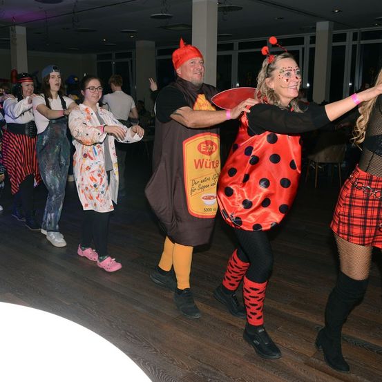 Eine Gruppe von Menschen tanzt auf einer Party. Eine Person ist als Marienkäfer verkleidet. Eine andere trägt ein Senfkostüm. Der Rest trägt Kostüme und Gesichtsbemalung. Der Raum ist schwach beleuchtet.