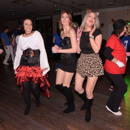 Eine Gruppe von Frauen tanzt auf einem Holzboden bei einer Party, in verschiedenen Kostümen. Eine Frau in Schwarz und Rot, eine andere in Schwarz und Leopardenmuster.