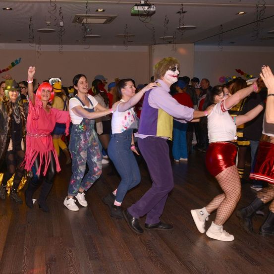 Eine Gruppe von Menschen tanzt auf einem Holzboden, einige in Kostümen. Ein Mann trägt Clown-Gesichtsfarbe und ein farbenfrohes Outfit. Andere tragen Stiefel, Brillen, Hüte und Armbänder. Ein Projektor und Luftballons sind im Hintergrund.