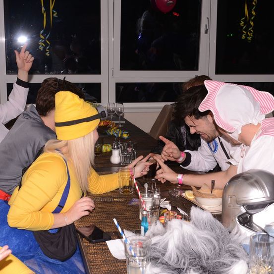 Eine Gruppe von Menschen in Kostümen sitzt um einen Tisch mit Essen und Getränken. Eine Frau in einem gelben Kostüm hält ein Getränk. Der Raum hat große Fenster und Dekorationen.