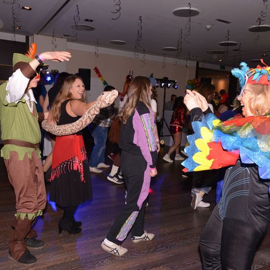 Menschen in Kostümen tanzen auf einer Party. Eine Frau in der Mitte trägt eine Schlangenattrappe. Der Raum hat einen Holzboden und Dekorationen.