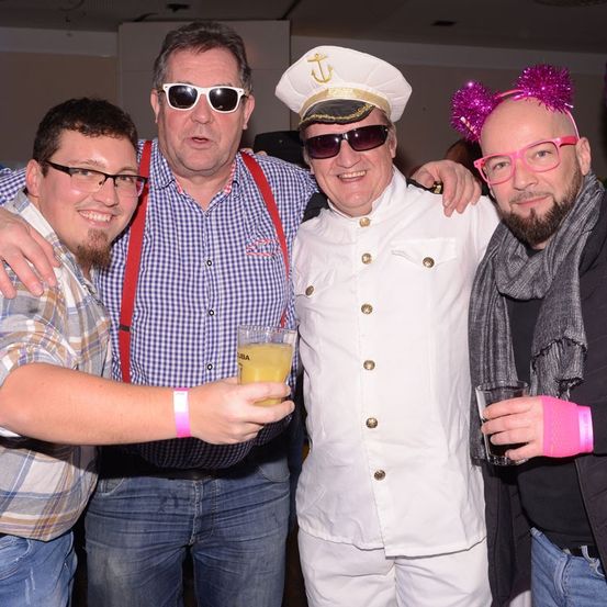 Vier Männer posieren für ein Foto. Ein Mann ist als Kapitän gekleidet, und ein anderer hat eine pinke Haarspange. Sie alle halten Getränke und lächeln.
