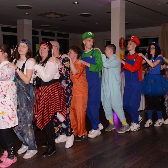 Eine Gruppe von Menschen in verschiedenen Kostümen, darunter Mario und Luigi, steht auf einer Tanzfläche. Sie tragen Turnschuhe und einige haben Armbänder an den Handgelenken. Im Hintergrund befinden sich Fenster und eine Decke mit Lichtern.