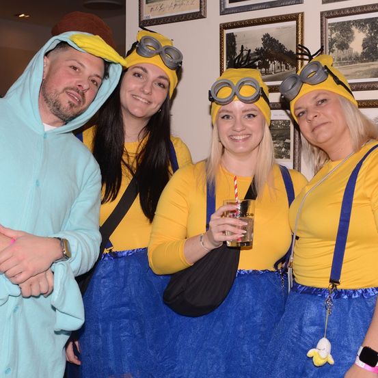 Vier Personen in gelben Kostümen mit Schutzbrillen und blauen Röcken. Der Mann auf der linken Seite trägt ein blaues Minion-Kostüm. Sie stehen eng beieinander und lächeln. Dahinter befinden sich gerahmte Bilder an der Wand.