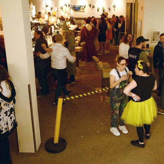Mehrere Menschen versammeln sich in einem Barbereich, einige in Kostümen. Eine Frau in einem gelben Tutu und ein Mann in einem schwarzen Shirt stehen sich gegenüber. Ein gelber Pfosten blockiert den Eingang.