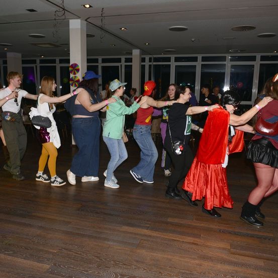 Eine Gruppe von Menschen in Kostümen tanzt auf einem Holzboden. Eine Frau in einem roten Umhang führt den Tanz an, während andere folgen. Der Raum hat große Fenster und eine Decke mit Lichtern.