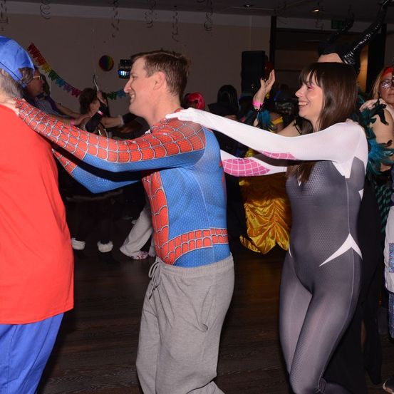 Eine Gruppe von Menschen tanzt auf einer Party. Ein Mann in Spider-Man-Kostüm und eine Frau in schwarz-weißem Outfit tanzen gemeinsam.