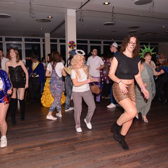 Eine Gruppe von Menschen in verschiedenen Kostümen tanzt auf einem hölzernen Boden in einer Einrichtung. Eine Frau trägt eine Statue der Freiheit als Kopfschmuck, während eine andere Frau einen Leoparden-Print-Rock trägt.
