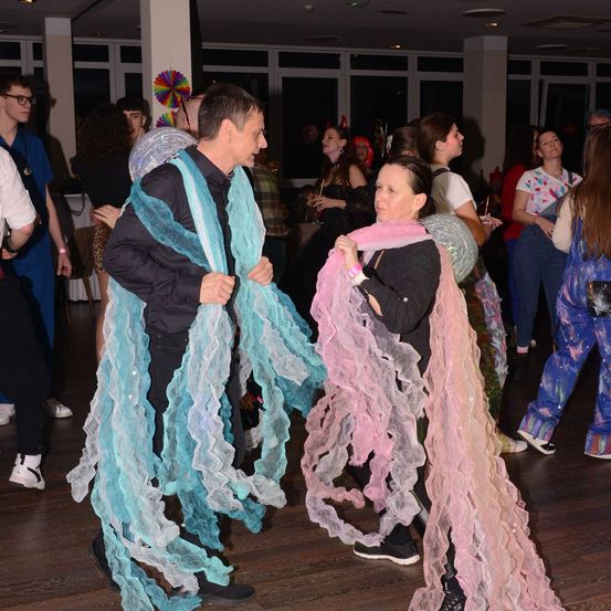 Eine Gruppe von Menschen tanzt in einem Raum. Zwei Personen mit rosa und blauen Umhängen sind im Vordergrund.