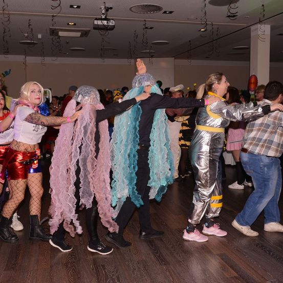 Eine Gruppe von Menschen tanzt auf einem Holzboden. Einige tragen Kostüme mit großen Rüschen und Helmen. Eine Person trägt ein silbernes Metallic-Outfit.