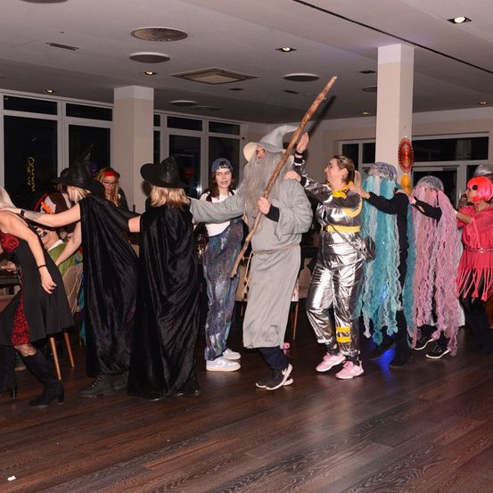 Eine Gruppe von Menschen in Kostümen, einschließlich Gandalf und Hexen, tanzt auf einem Holzboden in einem Raum mit Fenster und Lichtern.