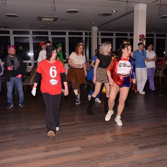 Mehrere Menschen in Kostümen tanzen auf einem Holzboden, während andere herumstehen und zusehen. Eine Frau in einem roten Kostüm mit der Nummer 6 geht in der Mitte.