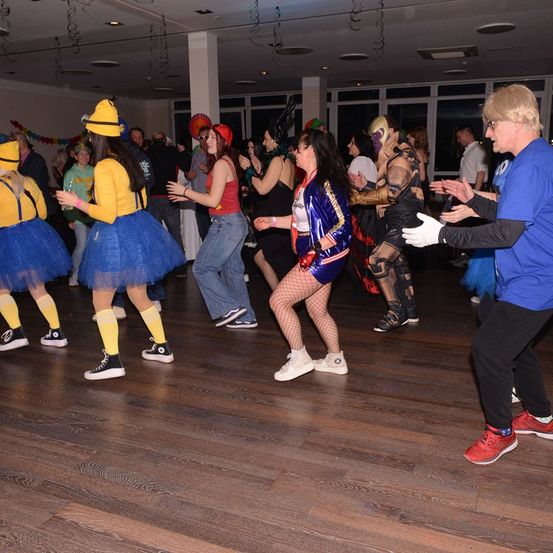 Eine Gruppe von Menschen in Kostümen tanzt in einem Saal. Ein Mann in Blau unterhält sich mit einer Frau in einem roten Kostüm. Eine weitere Person in einem gelben Kostüm steht in der Nähe.