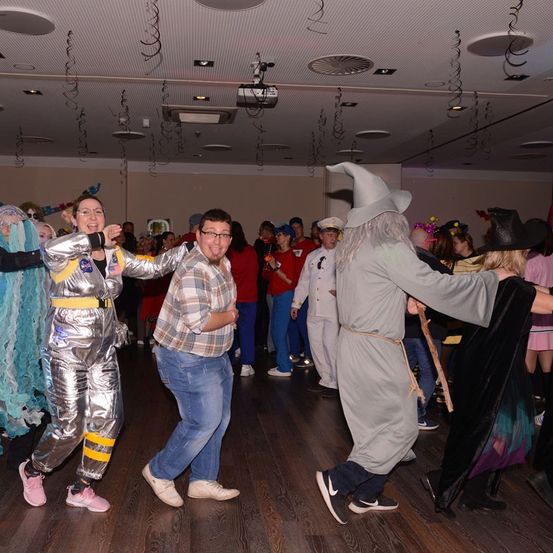 Eine Gruppe von Menschen tanzt in einem Raum mit Holzböden, einige sind in Kostümen gekleidet. Ein Mann in einem Zaubererhut tanzt mit einer Frau in einer silbernen Anzug.