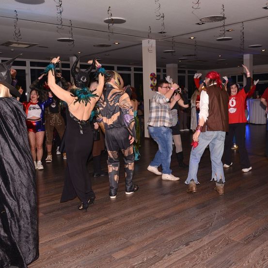 Eine Gruppe von Menschen tanzt in einem Saal, trägt verschiedene Kostüme, einige in hohen Absätzen und andere in Stiefeln. Sie sind von Lichtern beleuchtet.