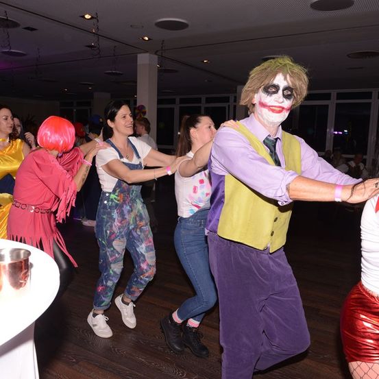 Eine Gruppe von Menschen in Kostümen tanzt in einem Saal. Ein Mann als Joker hält zwei Frauen im Kreis.