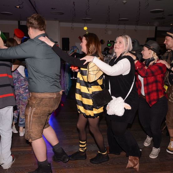Mehrere Menschen tanzen auf einem Holzboden in einem Partyraum. Eine Frau in einem Bienenkostüm hält eine andere Frau beim Tanzen. Ein Mann in traditioneller Kleidung hält eine Frau mit Hut.