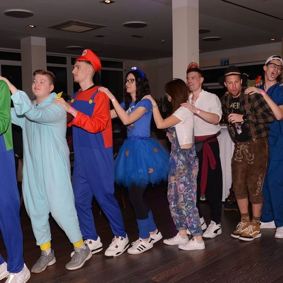 Eine Gruppe von Menschen in Kostümen tanzt auf einem Holzboden. Sie tragen Turnschuhe und einige tragen Armbänder. Die Leute halten sich an den Händen und scheinen Spaß zu haben. Der Raum hat eine weiße Säule und Glasfenster.