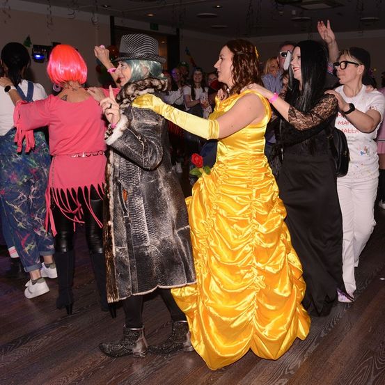 Mehrere Menschen tanzen in einem Partyraum. Eine Frau in einem gelben Kleid tanzt mit zwei anderen. Eine Frau in Pink tanzt mit einer anderen Person.