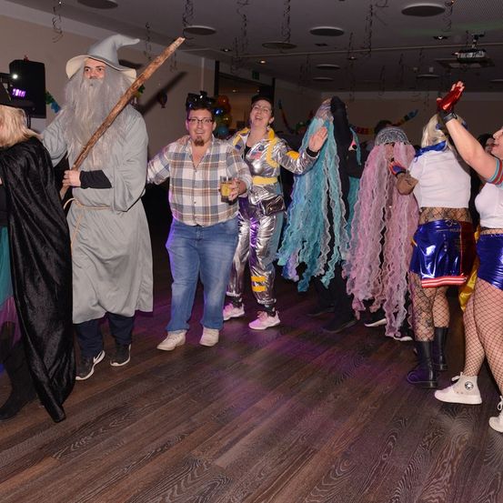 Mehrere Menschen sind in verschiedenen Kostümen, eines als Gandalf, ein anderes als Raumanzug, auf einer Party, tanzen und lächeln.