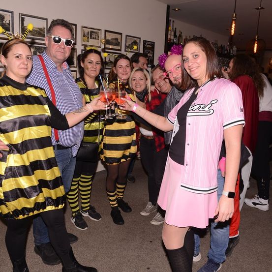 Eine Gruppe von Erwachsenen in Kostümen posiert für ein Foto in einer Bar, hält Getränke in der Hand und trägt Brillen. Die Frau auf der rechten Seite trägt ein pinkes Shirt mit Text. Der Mann auf der linken Seite hat Hosenträger.