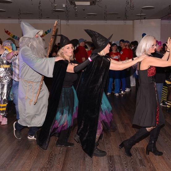 Ein Mann und zwei Frauen in Kostümen tanzen auf einem Holzboden zusammen, umgeben von anderen Menschen in Kostümen.