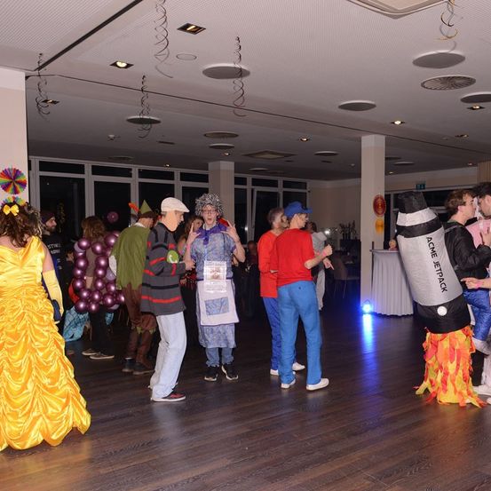 Eine Gruppe von Menschen in Kostümen steht und unterhält sich in einem Raum mit Holzboden. Eine Person trägt ein gelbes Kleid und Handschuhe.