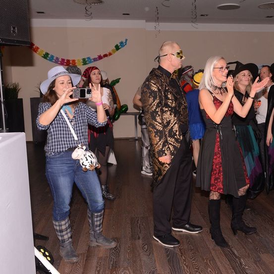 Eine Gruppe von Menschen in einem Partysaal. Eine Frau macht ein Foto mit ihrem Handy. Andere tragen Kostüme und Hüte. Der Raum hat Dekorationen und einen Holzboden.