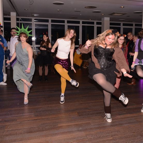 Eine Gruppe von Frauen tanzt in einem Partysaal, einige in Kostümen. Eine Frau trägt eine Statue of Liberty-Kopfbedeckung. Der Boden ist aus Holz und glänzend.